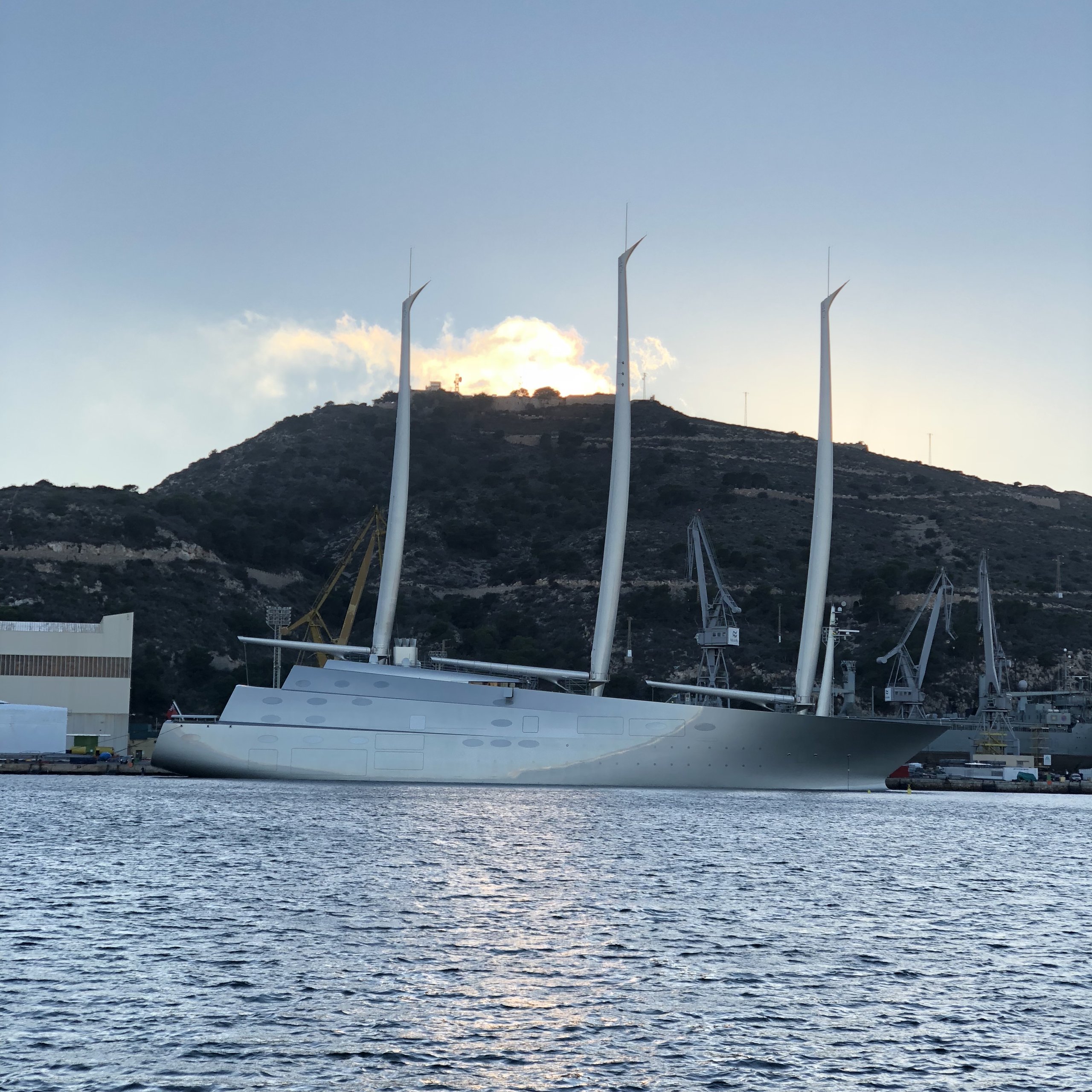 A vast three-masted sailing yacht moored at dusk, harbour cranes behind